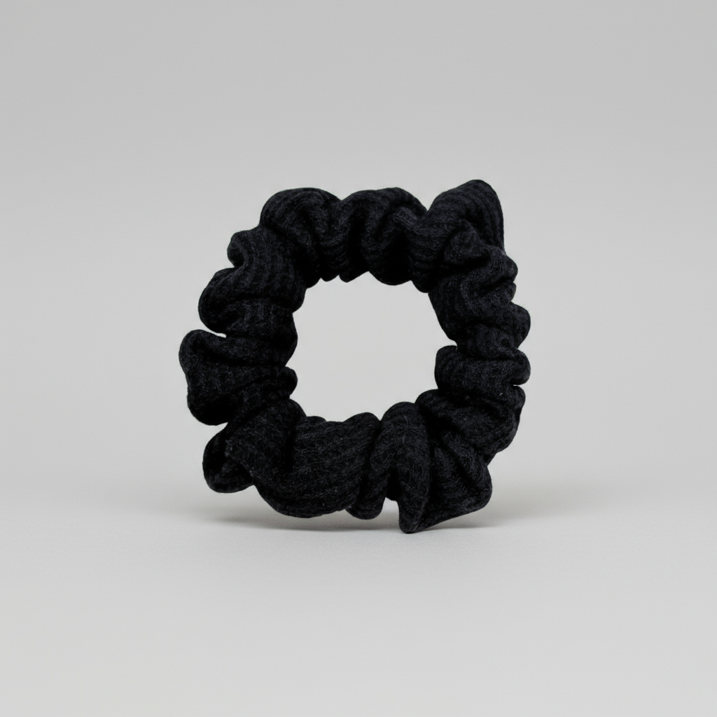 Black waffle knit cotton scrunchie with a gathered, ruffled texture, displayed against a neutral background. The ribbed fabric adds subtle dimension to the sleek black tone, offering a soft, secure hold ideal for everyday styling.