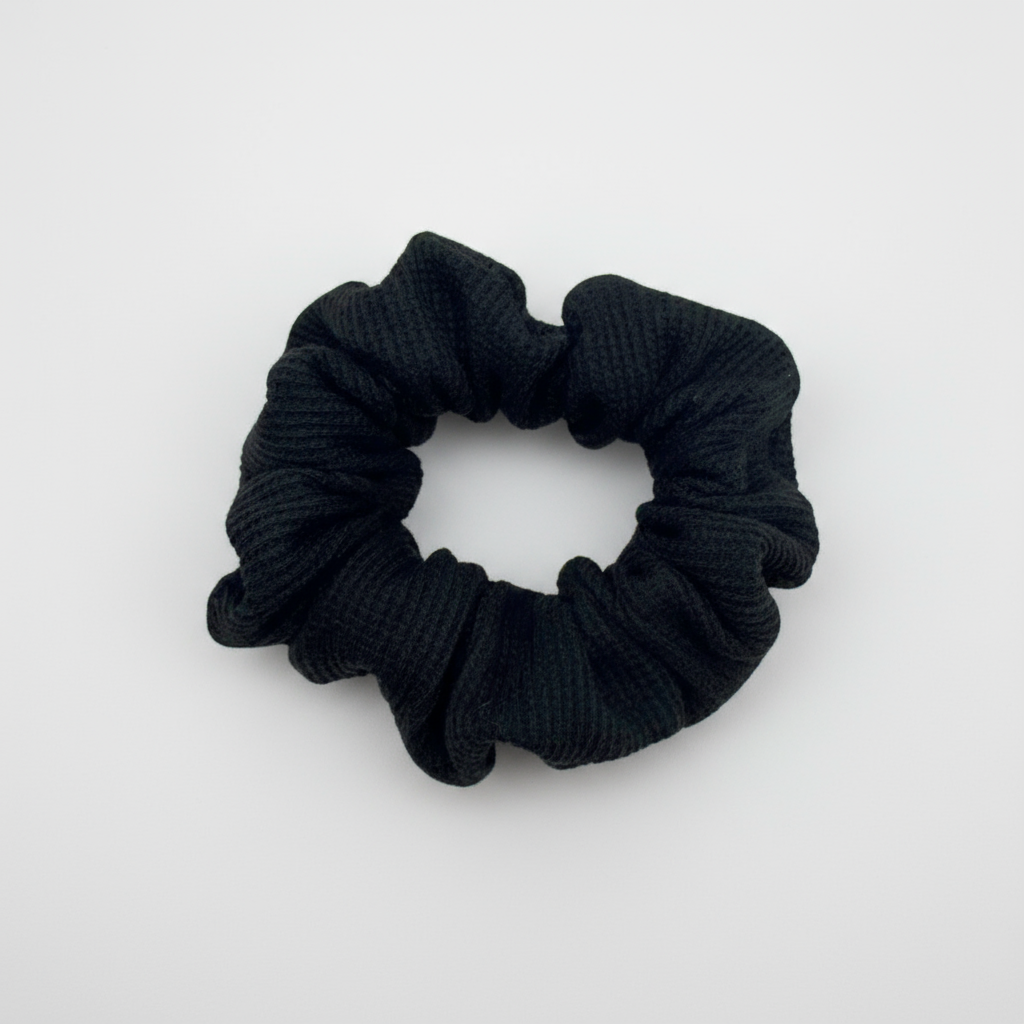 Black waffle knit cotton scrunchie with a gathered, ruffled texture, displayed against a neutral background. The ribbed fabric adds subtle dimension to the sleek black tone, offering a soft, secure hold ideal for everyday styling.