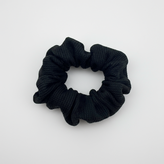 Black waffle knit cotton scrunchie with a gathered, ruffled texture, displayed against a neutral background. The ribbed fabric adds subtle dimension to the sleek black tone, offering a soft, secure hold ideal for everyday styling.